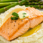 A close-up of perfectly seared salmon topped with melted butter, resting on creamy mashed potatoes and paired with asparagus.